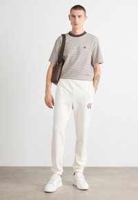 Pasiasty, krótki rękaw T-shirt w kolorze brązowym i białym, zestawiony z białymi spodniami dresowymi z czerwonym logo. W zestawie białe sneakersy i brązowa torba.