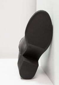 Zapato de cuero negro con suela de goma texturizada, inclinado contra un fondo blanco. Presenta un tacón cuadrado y un diseño suave y redondeado.