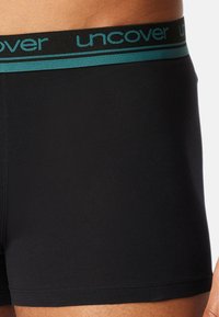 Black boxer briefs with a contrasting turquoise waistband featuring "uncover" text. Smooth, elastic fabric for comfort, mid-thigh length.