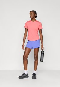 Ροζ αθλητικό μπλουζάκι Nike, ελαφρύ ύφασμα, συνδυασμένο με μπλε σορτς. Το μοντέλο κρατάει μαύρο παγούρι, φοράει μαύρα αθλητικά παπούτσια και λευκές κάλτσες.