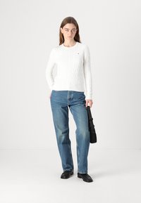 Pull en tricot blanc, jean bleu, chaussures noires et sac à bandoulière noir. Le pull présente des poignets et un col côtelés ; le jean a une coupe décontractée.