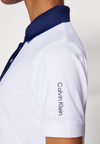 Bílá polokošile s námořnickým límcem a manžetami, s nápisem "Calvin Klein" vytištěným v námořnické barvě na pravém rukávu. Hladká textilie.