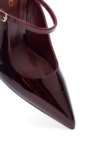 Zapato Mary Jane de charol burdeos con punta afilada, tirante delgado al tobillo y hebilla dorada sobre fondo blanco.