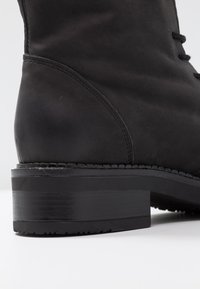Bottine en cuir noir avec une finition lisse, bout rond et coutures surpiquées. Elle présente un talon carré et un laçage à l'avant.