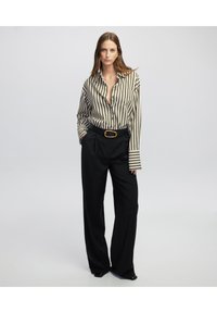 Pantaloni neri a gamba larga abbinati a una camicia con bottoni a righe verde e bianco. La camicia presenta maniche lunghe e una tasca sul petto, con una cintura dorata.