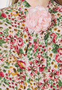 Blouse florale avec un fond rose, des feuilles vertes et des fleurs rouges, accentuée par une grande fleur en satin rose au niveau du col.