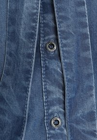 Camicia di jeans blu con una texture morbida, con un evidente dettaglio di bottoni al collo e cuciture sottili lungo la parte anteriore.