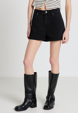 Donna che indossa pantaloncini di jeans neri a vita alta, top corto a righe e stivali di pelle neri al ginocchio, in piedi su uno sfondo semplice.