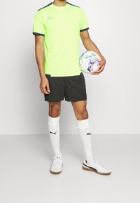 Camiseta deportiva verde neón con detalles en negro, combinada con pantalones cortos negros y calcetas blancas. Sosteniendo un balón de fútbol colorido.