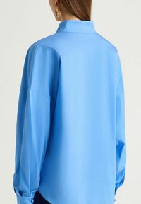 Camicia blu con colletto alto, vestibilità morbida e maniche lunghe leggermente a sbuffo con dettagli a bottone. Tessuto liscio e colore uniforme.