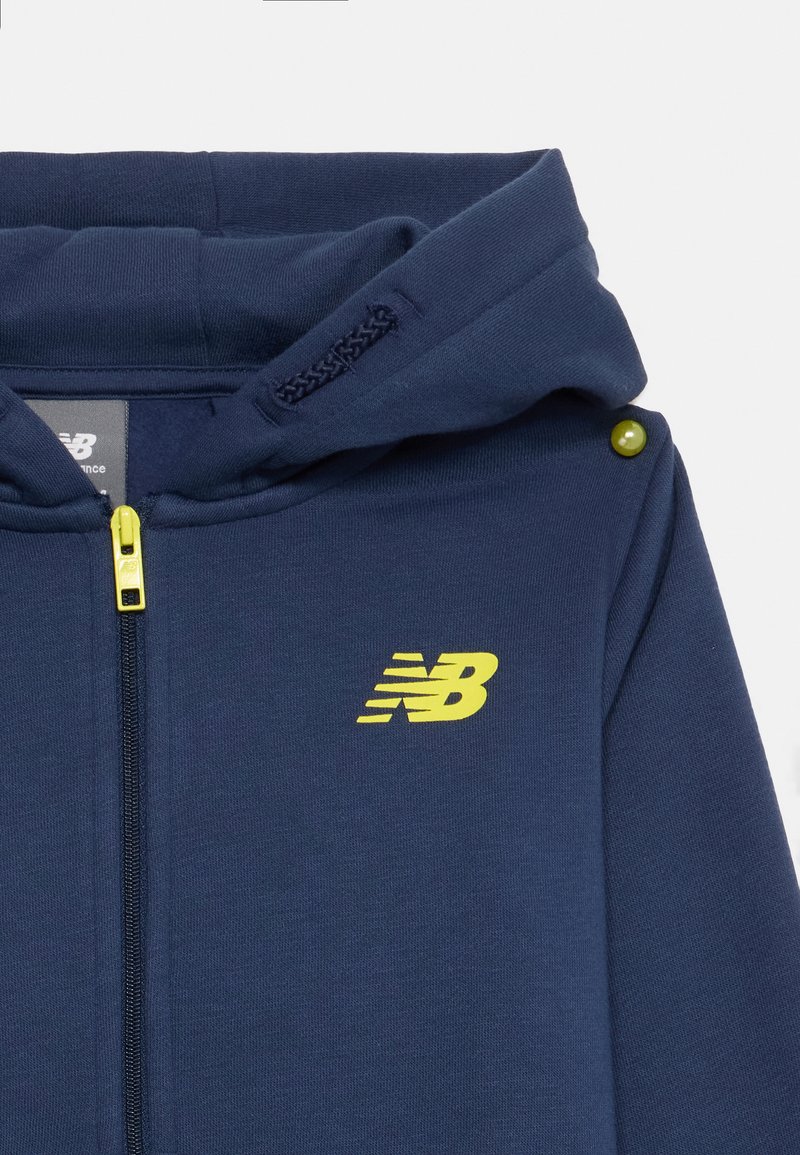 Felpa con zip di colore blu navy realizzata in tessuto morbido, con logo New Balance giallo, chiusura a zip e cappuccio staccabile con hardware verde.