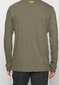 Achteraanzicht van een persoon in een olijfgroene langen mouwen Under Armour-shirt en zwarte sportbroek met witte zijstrepen.