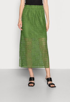 Femme portant une jupe en dentelle verte longueur genou avec des motifs floraux carrés, associée à des sandales noires à talons et un haut noir.