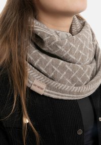 Beige gebreide infinity-sjaal met een geometrisch patroon en een gebreide textuur, voorzien van een stoffen label met "Knit Factory."