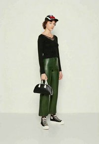 Kvinne som har på seg svart langermet topp, grønne skinnbukser, svart og hvit joggesko, svart og hvit caps, og holder en svart veske med hvite håndtak.