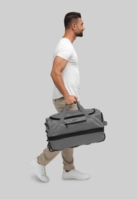 Borsa da viaggio grigia con scomparti con cerniera, manici robusti e superficie liscia. Utente che trasporta la borsa, indossando abbigliamento leggero e scarpe da ginnastica.