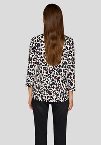 Woman with long brown hair wearing a white blouse with a black, red, and purple abstract pattern and black pants, facing away from the camera.