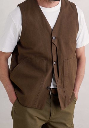 Mann trägt eine braune, geknöpfte Weste über einem weißen kurzärmligen T-Shirt mit den Händen in den Taschen olivgrüner Hosen.