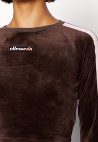 Ruskea samettinen pitkähihainen paita, jossa on pyöreä pääntie. Olkapäissä vaaleanpunaiset raidat ja rinnassa musta kirjailtu "ellesse" -logo.
