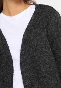 Grafitowo-szary, teksturowany kardigan o luźnym, otwartym kroju, noszony na białej koszuli, ukazujący puszyste włókna i swobodną sylwetkę.