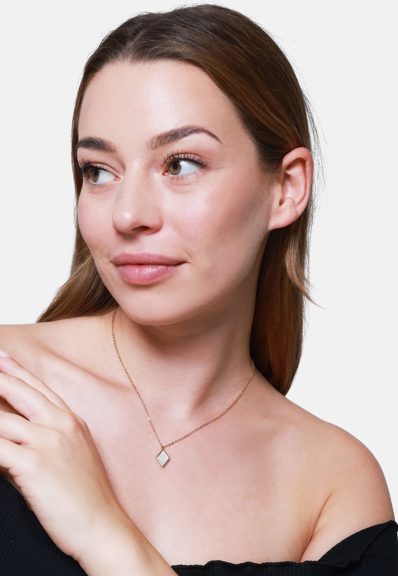 Goldkette mit einem diamantförmigen Anhänger mit reflektierender Oberfläche, der auf nackter Haut getragen wird. Strukturiertem Stoff des schulterfreien Oberteils sichtbar.