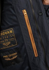 Marineblaues Jacket mit strukturiertem Innenfutter, orangefarbener Reißverschlussöffnung und Branding-Labels in Gold auf schwarzem Stoff, das ein praktisches Design zeigt.