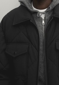 Persoon in gelaagde kleding met een zwart gewatteerd jack over een grijze gebreide hoodie die dichtgeritst is, en een witte ronde hals T-shirt eronder.