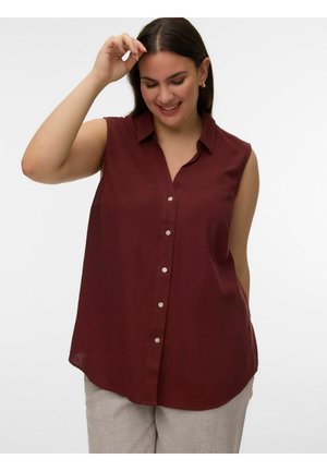 Femeie zâmbitoare, îmbrăcată într-o cămașă fără mâneci bordo cu nasturi și pantaloni bej deschis, ridicând o mână lângă cap pe un fundal alb simplu.