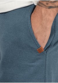 Blue garment with a soft texture, featuring a side pocket and a brown leather accent tag. Smooth fabric with a contemporary design.