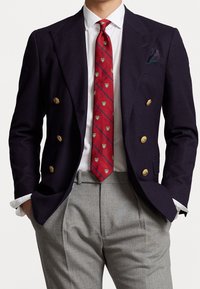 Męska dwurzędowa granatowa marynarka z złotymi guzikami, biała koszula i czerwony krawat z wzorzystymi motywami tarczy, w połączeniu z szarymi spodniami.