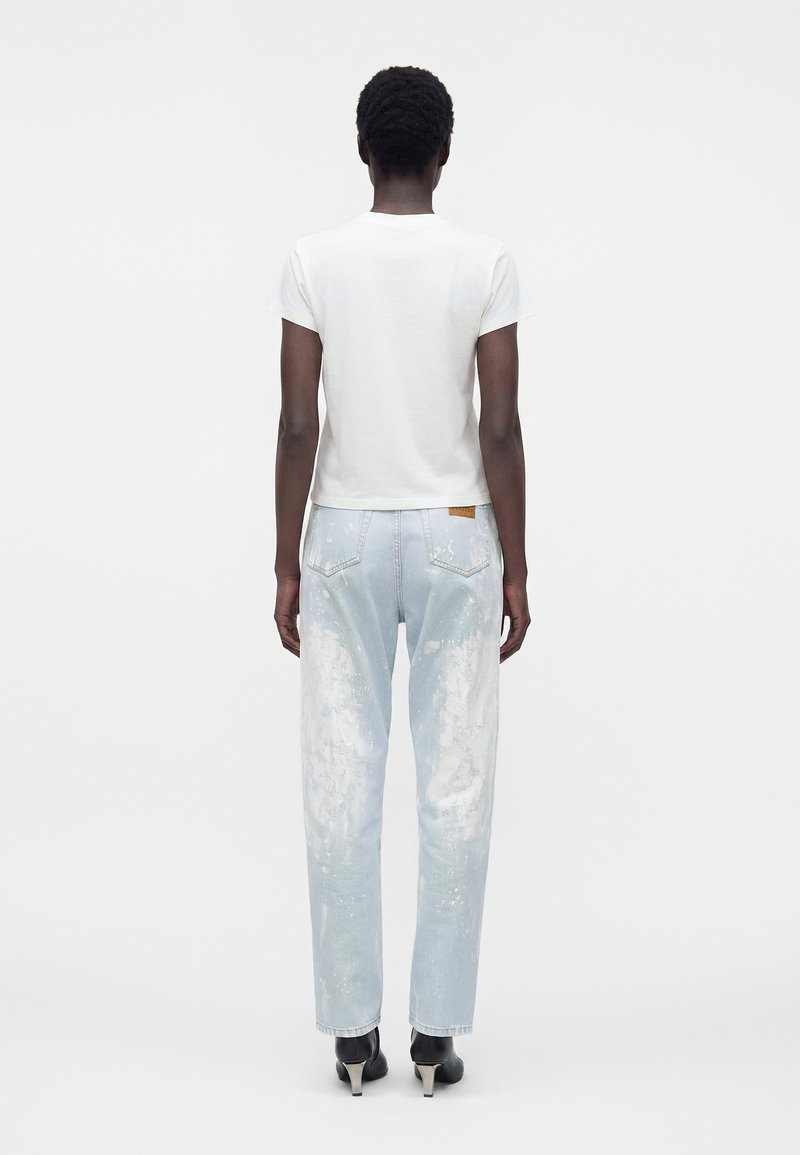 Person standing facing away wearing a white short-sleeve shirt, light blue splattered jeans, and black heeled ankle boots.