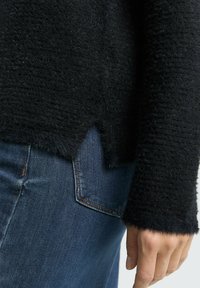 Black fuzzy sweater with a textured pattern, side slit, and ribbed cuffs, paired with blue denim jeans featuring a visible back pocket.