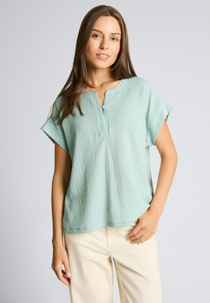 Młoda kobieta o długich brązowych włosach, ubrana w teksturowaną jasnoniebieską bluzkę z krótkim rękawem i kremowe spodnie, stojąca na jednolitym tle.