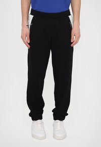 Sorte joggingbukser med slim fit, der har sidelommer med lynlås og ribbet tekstur på sidepanelerne, parret med hvide sneakers.