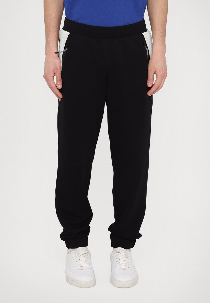Sorte joggingbukser med slim fit, der har sidelommer med lynlås og ribbet tekstur på sidepanelerne, parret med hvide sneakers.