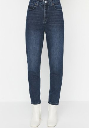 Donkerblauwe high-waist slim-fit jeans gecombineerd met witte enkellaarzen, getoond op een witte achtergrond, bij de taille en enkels bijgesneden.