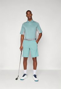 Lys blå golfpolo med grå detaljer, kombinert med matchende shorts. Modellen holder en golfkølle og har på seg hvite sko og sokker.