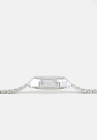 Montre en metal argenté avec un boîtier rectangulaire, un bracelet texturé blanc et deux boutons-poussoirs sur le côté. Le verre plat affiche l'heure.