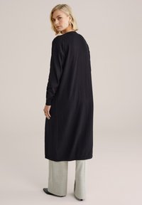 Abito lungo nero in tessuto morbido con maniche lunghe e scollo arrotondato, abbinato a pantaloni a gamba larga grigio chiaro e tacchi neri.