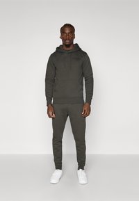 Donkergroene hoodie en bijpassende taps toelopende broek van fleece materiaal, met een voorzak en een trekkoord in de capuchon, gecombineerd met witte sneakers.