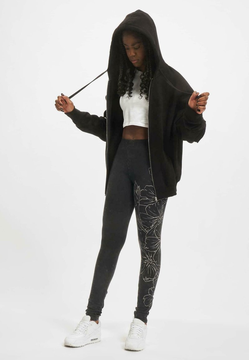 Svart hettejakke med glidelås og stor hette, hvit cropped skjorte under, svarte leggings med hvite blomsterprint, og hvite joggesko.