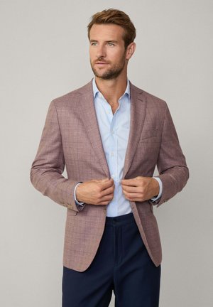 Blazer de cuadros en rosa claro con solapas de muesca y botones en tono dorado, combinado con una camisa de vestir azul clara, llevada abierta sobre pantalones marinos.