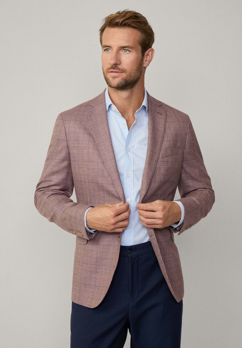 Blazer à carreaux rose clair avec des revers et des boutons dorés, associé à une chemise habillée bleu clair, portée ouverte sur un pantalon marine.