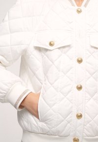 White quilted jacket featuring gold buttons, two chest pockets, and ribbed cuffs with a metallic accent. Soft texture and structured fit.