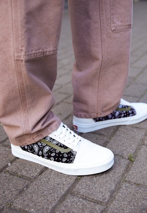 Fødder iført hvide sneakers med sort paisley-mønster og olivengrin, stående på grå teglstensbelægning, med lysebrune bukser.