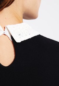 Svart stickad topp med en vit spetskrage och knäppning bak, som visar kontrasterande texturer och färger i en figurnära design.