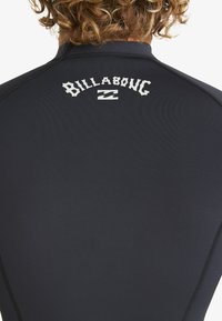 Czarny piankowy strój kąpielowy o gładkiej, fakturowanej powierzchni, z białym logo "Billabong" i wzorem fali na górnej części dekoltu z tyłu.