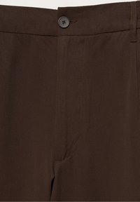Gros plan d'un pantalon brun foncé montrant la taille avec un bouton noir à quatre trous et une couture visible à l'avant.