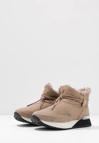Un par de botas de tobillo beige con forro suave, cordones envolventes, suelas de cuña en blanco y negro, y material texturizado sobre una superficie blanca.