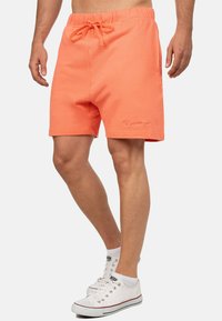 Koraalkleurige shorts van zacht materiaal, met een elastische tailleband en trekkoord, en een klein logo op het been. Witte sneakers worden weergegeven.